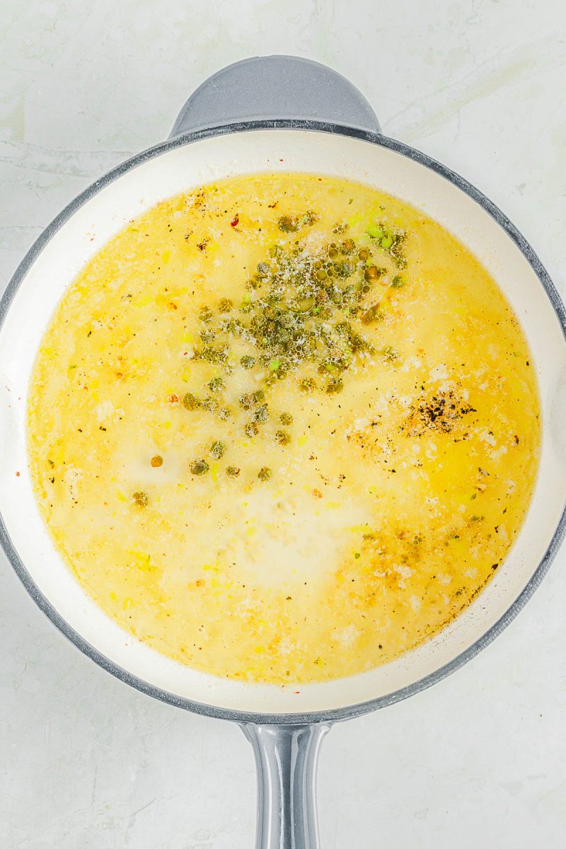 A white pot filled with a yellowish broth, herbs, and spices, sitting on a light-colored surface.