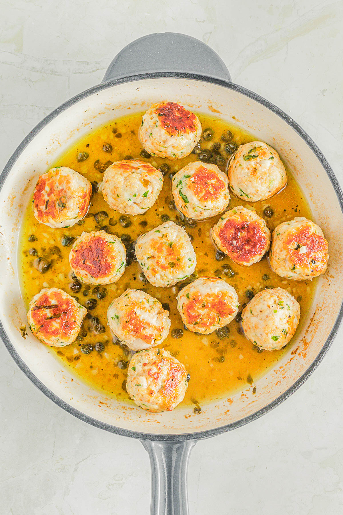 A skillet filled with browned meatballs in a yellow sauce with capers, viewed from above on a light surface.
