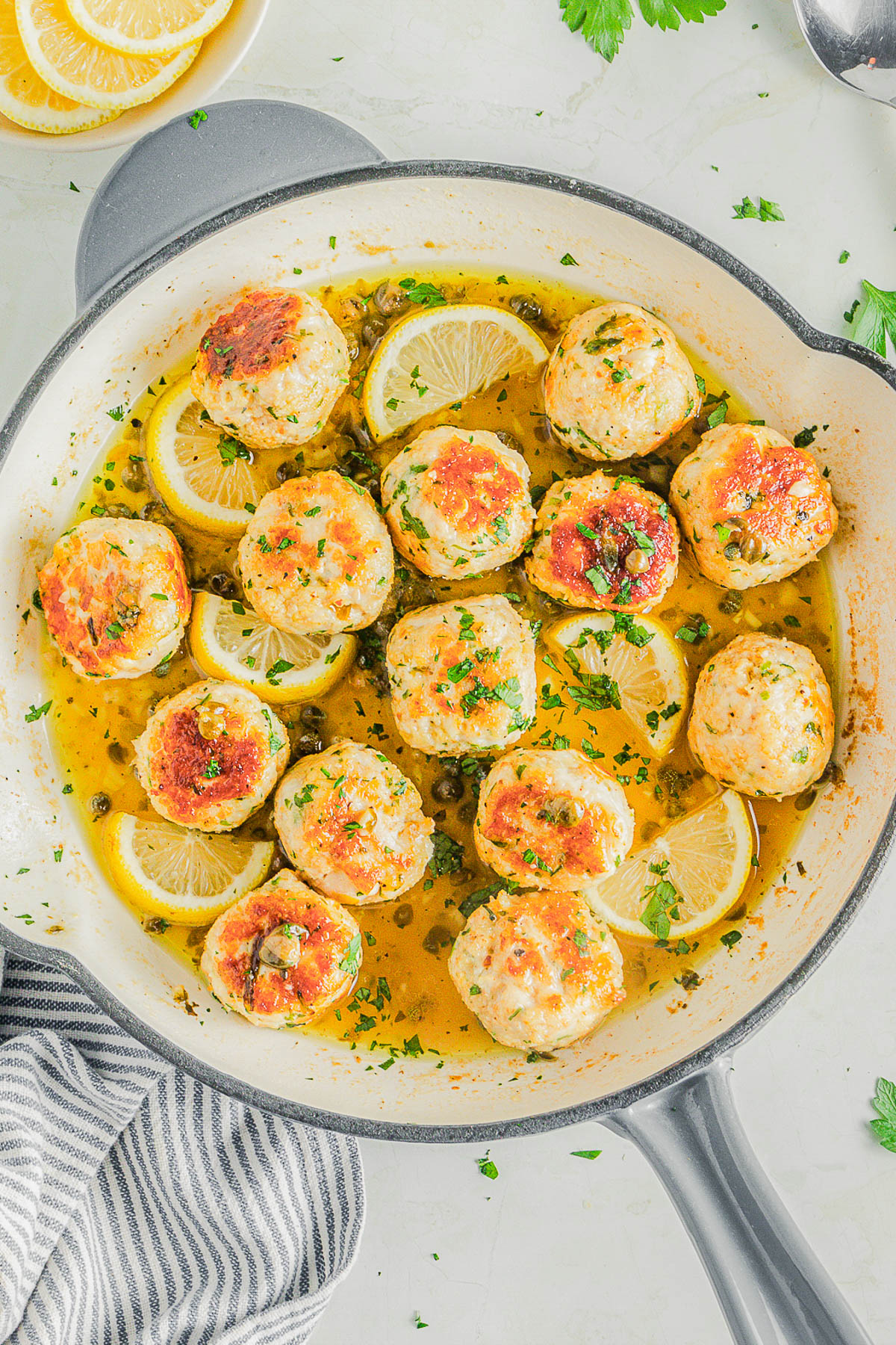 Chicken meatballs in a skillet with lemon slices, capers, and fresh herbs in a light sauce, viewed from above.