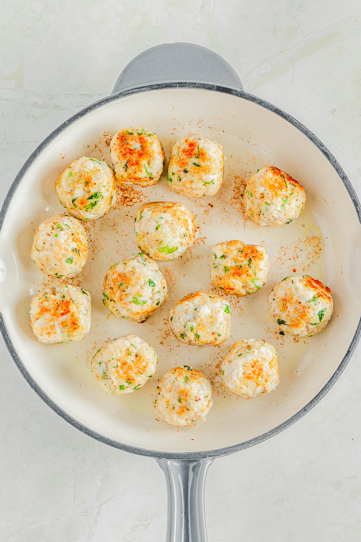 A white skillet with browned chicken meatballs containing green herbs, evenly spaced and cooked on a light-colored stovetop.