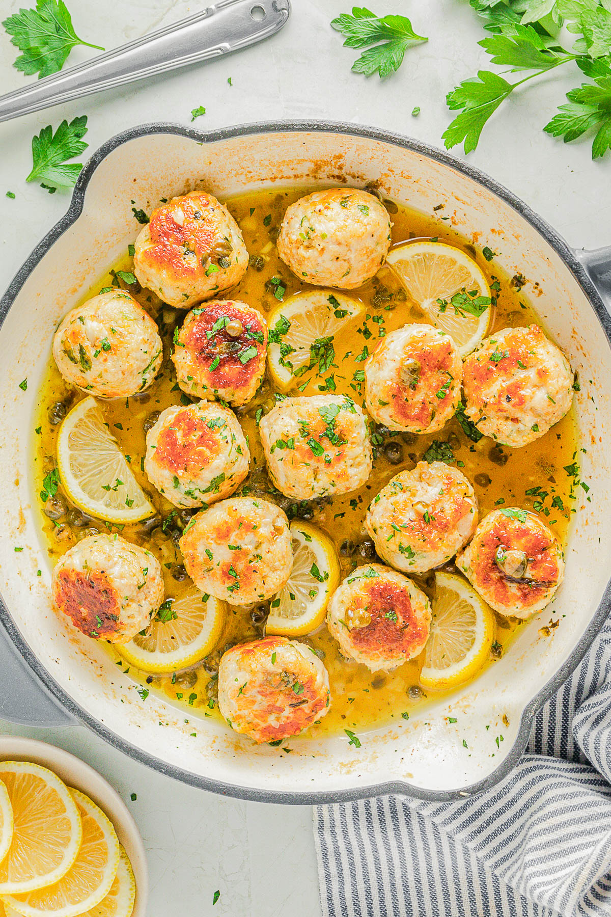 A skillet filled with browned chicken meatballs in a lemon sauce, garnished with lemon slices and chopped parsley.