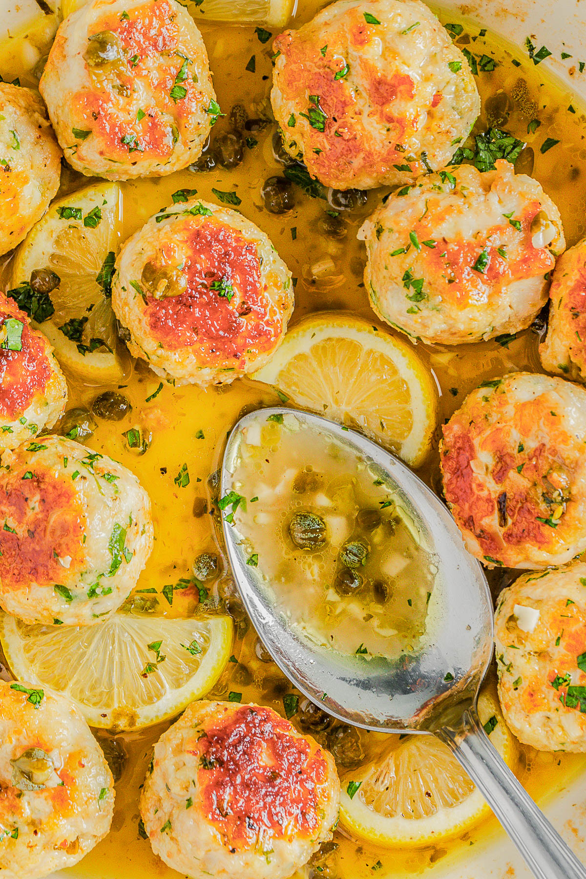 Chicken meatballs garnished with herbs in a lemon and caper sauce, surrounded by lemon slices, with a silver spoon resting in the dish.
