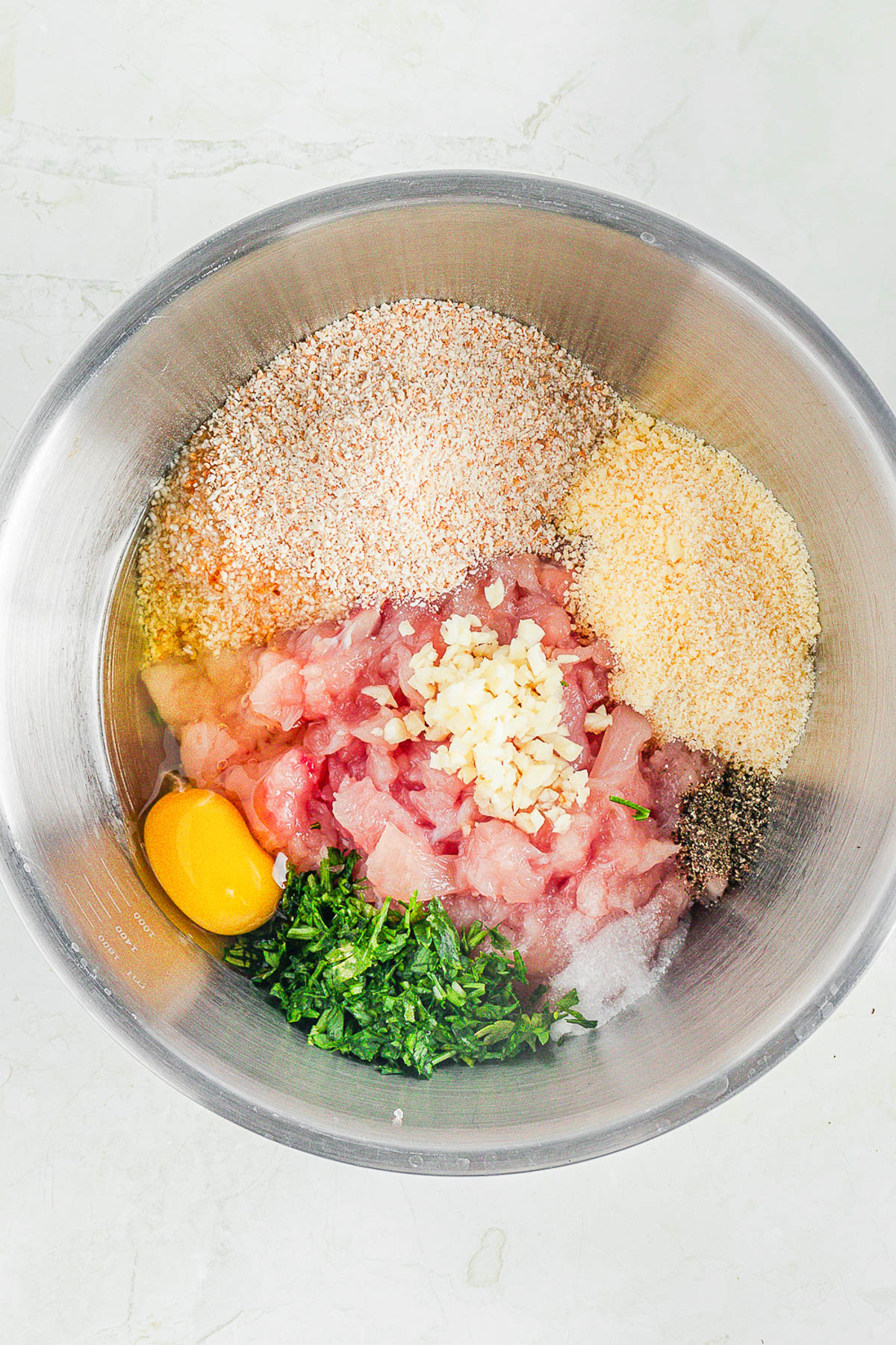 A metal bowl containing raw ground chicken, an egg, chopped parsley, minced garlic, breadcrumbs, salt, pepper, and grated cheese, ready for mixing.