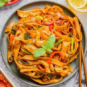 A plate of stir-fried flat rice noodles with chicken, bell peppers, basil, and sauce, garnished with fresh basil, with a drink, lime slices, and chili sauce on the side.