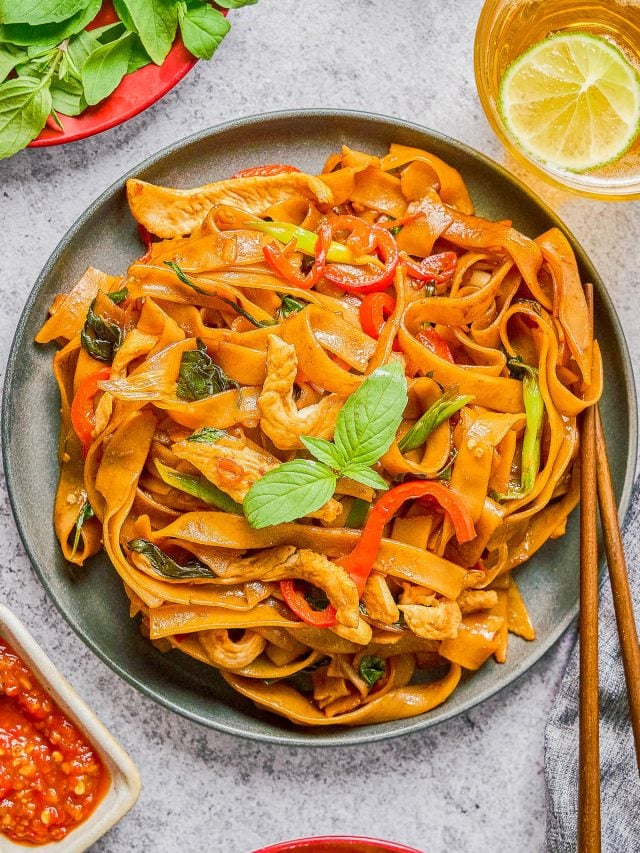 A plate of stir-fried flat rice noodles with chicken, bell peppers, basil, and sauce, garnished with fresh basil, with a drink, lime slices, and chili sauce on the side.