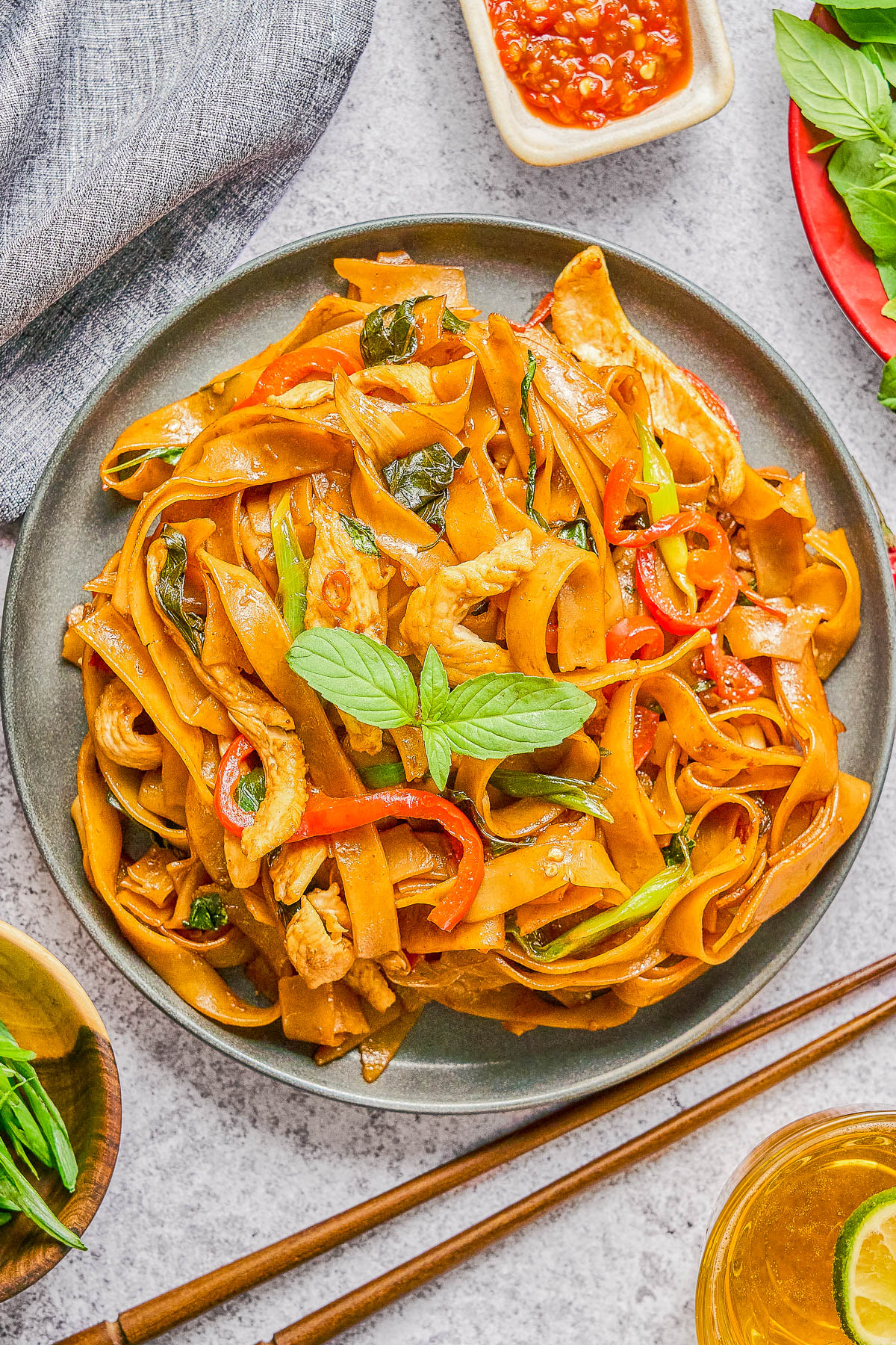 A plate of stir-fried wide rice noodles with chicken, bell peppers, green onions, and basil, garnished with fresh basil leaves. Chopsticks and sauces are placed nearby.