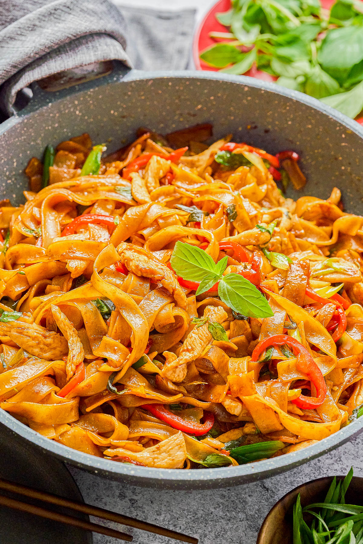 A pan filled with stir-fried flat noodles, sliced chicken, red bell peppers, green onions, and fresh basil leaves.