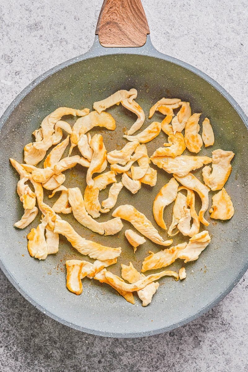 Sliced pieces of chicken breast are cooking in a gray non-stick frying pan on a light-colored countertop.