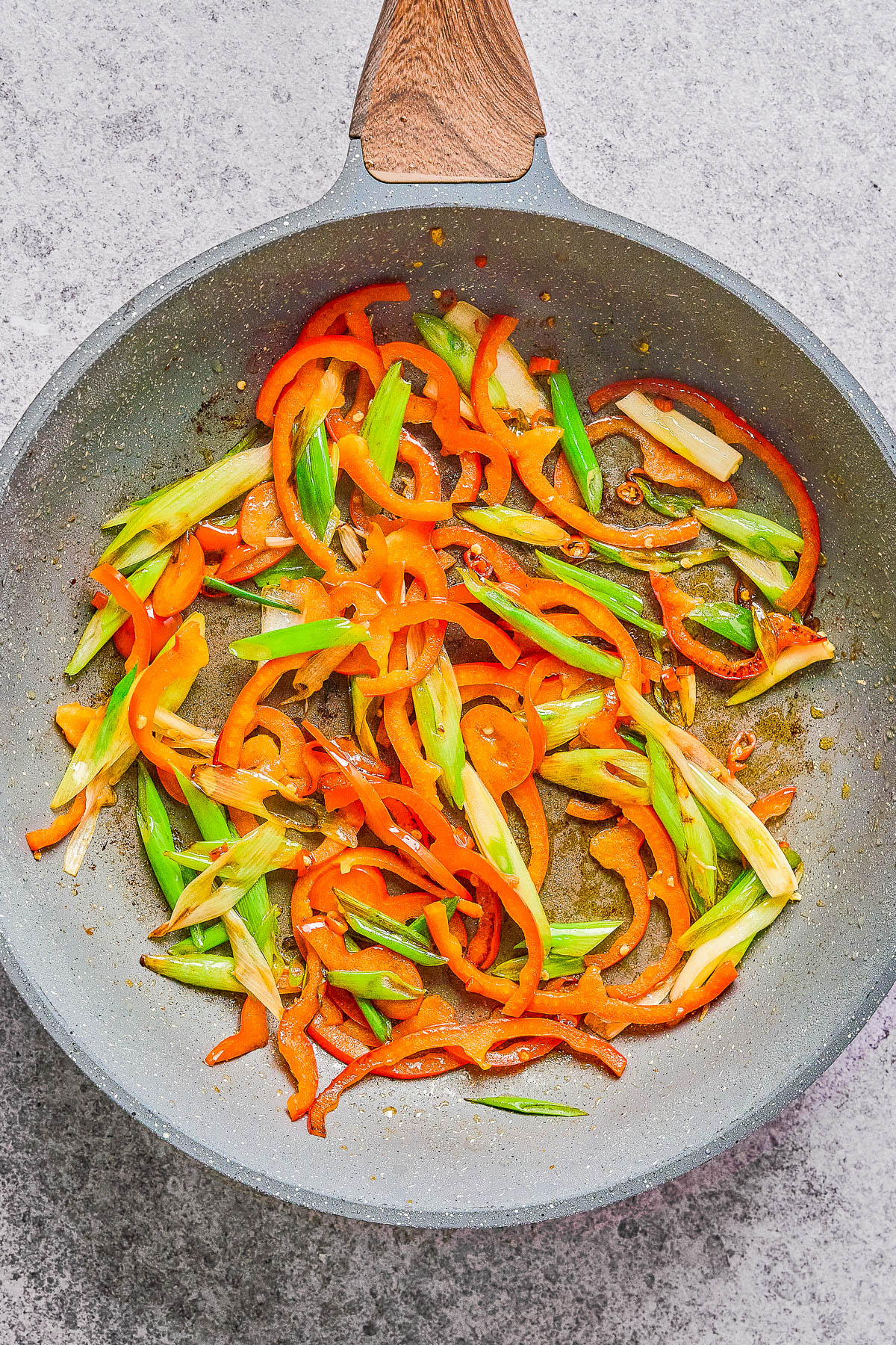 Sliced red bell peppers and green onions being sautéed in a nonstick skillet on a gray countertop.