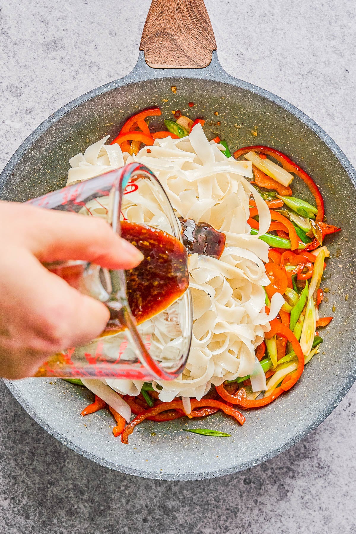 A hand pours sauce from a measuring cup into a pan containing rice noodles, sliced red bell peppers, and green onions.