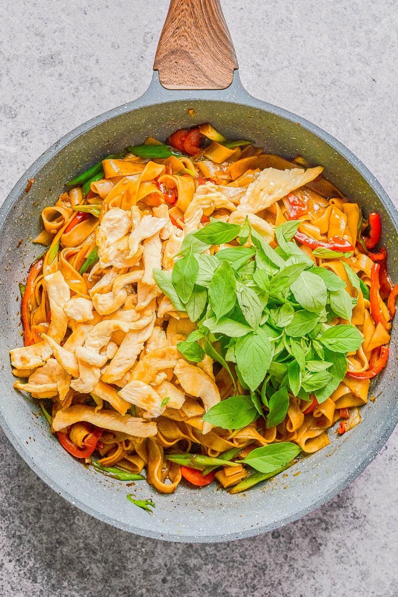 A skillet contains stir-fried noodles with sliced chicken, red bell peppers, green vegetables, and a handful of fresh basil leaves on top.