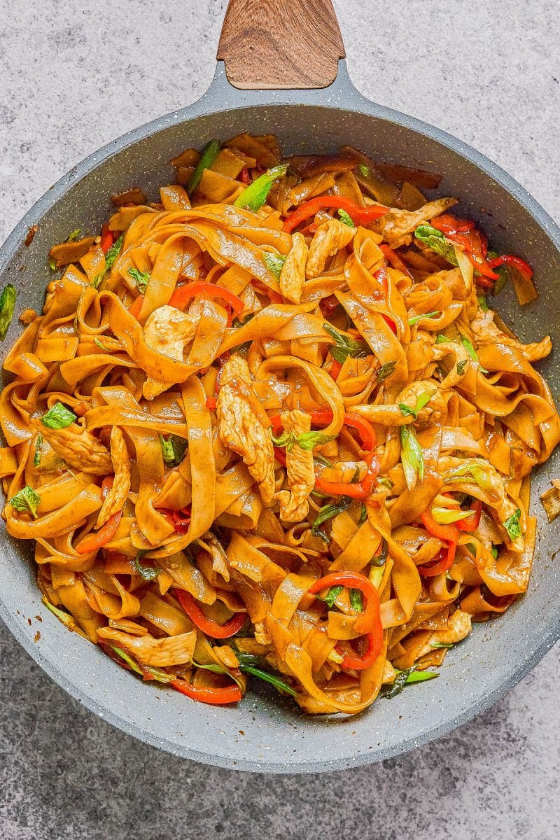 A pan filled with stir-fried rice noodles, sliced chicken, red bell peppers, and greens, all mixed in a brown sauce.