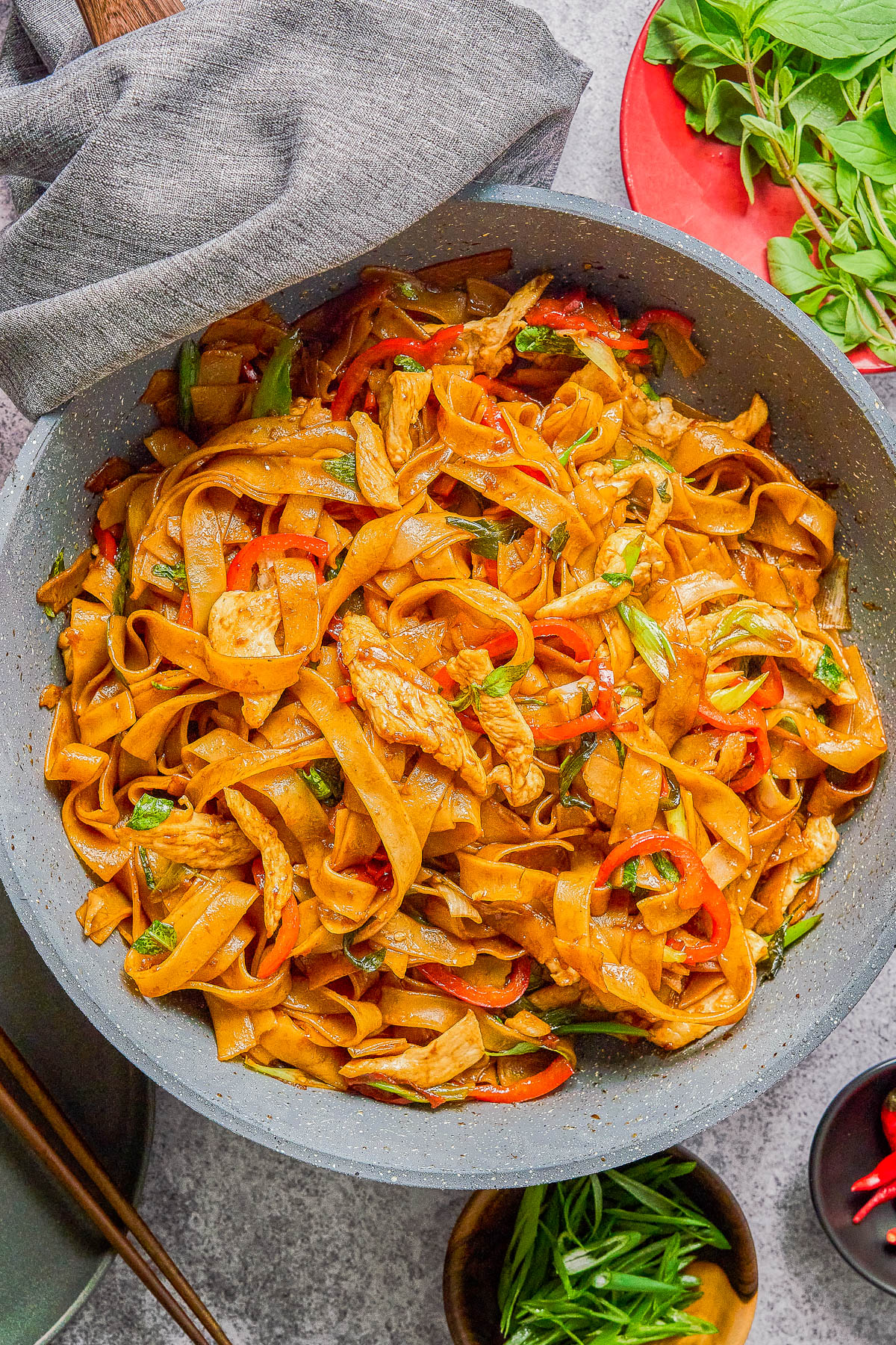 A wok filled with stir-fried flat rice noodles, sliced chicken, red bell peppers, and green herbs, with chopsticks and ingredient bowls nearby.
