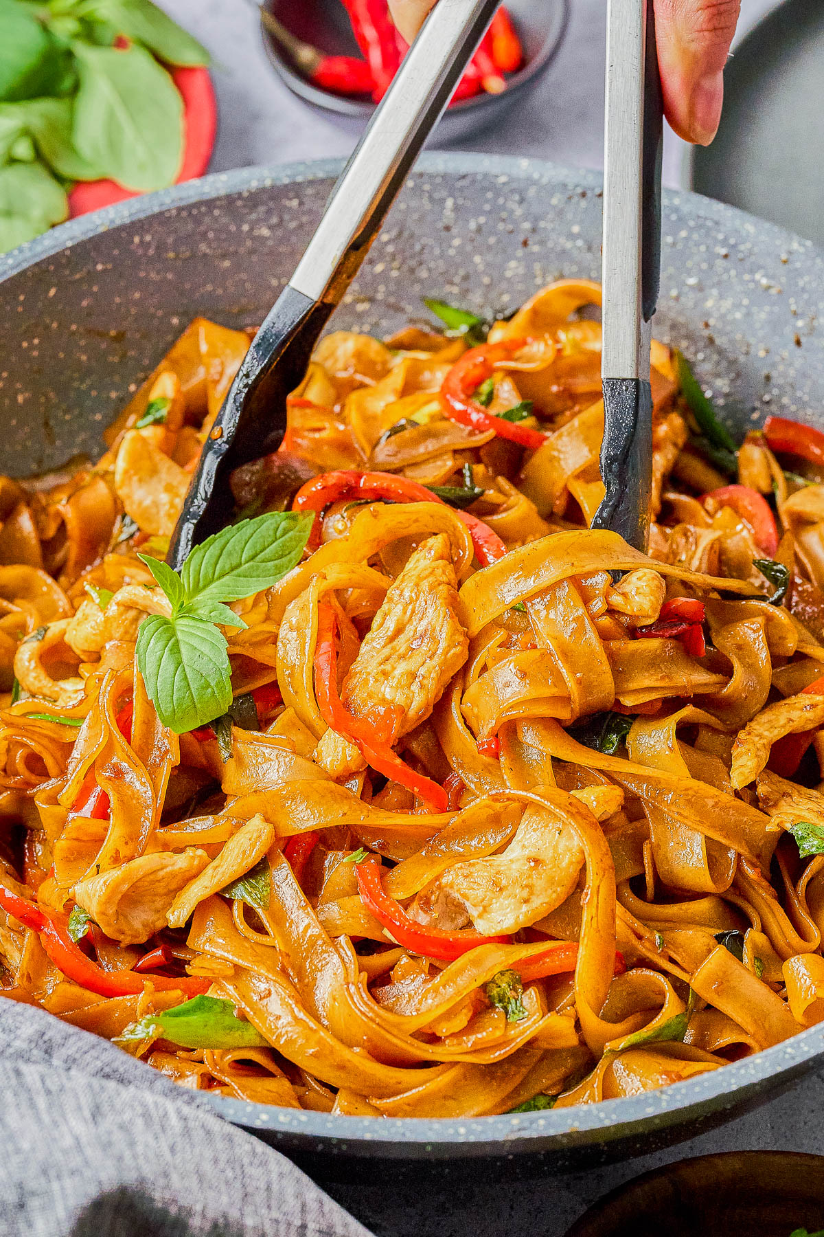 A pan of stir-fried noodles with sliced chicken, red bell peppers, and a garnish of basil, being mixed with tongs.