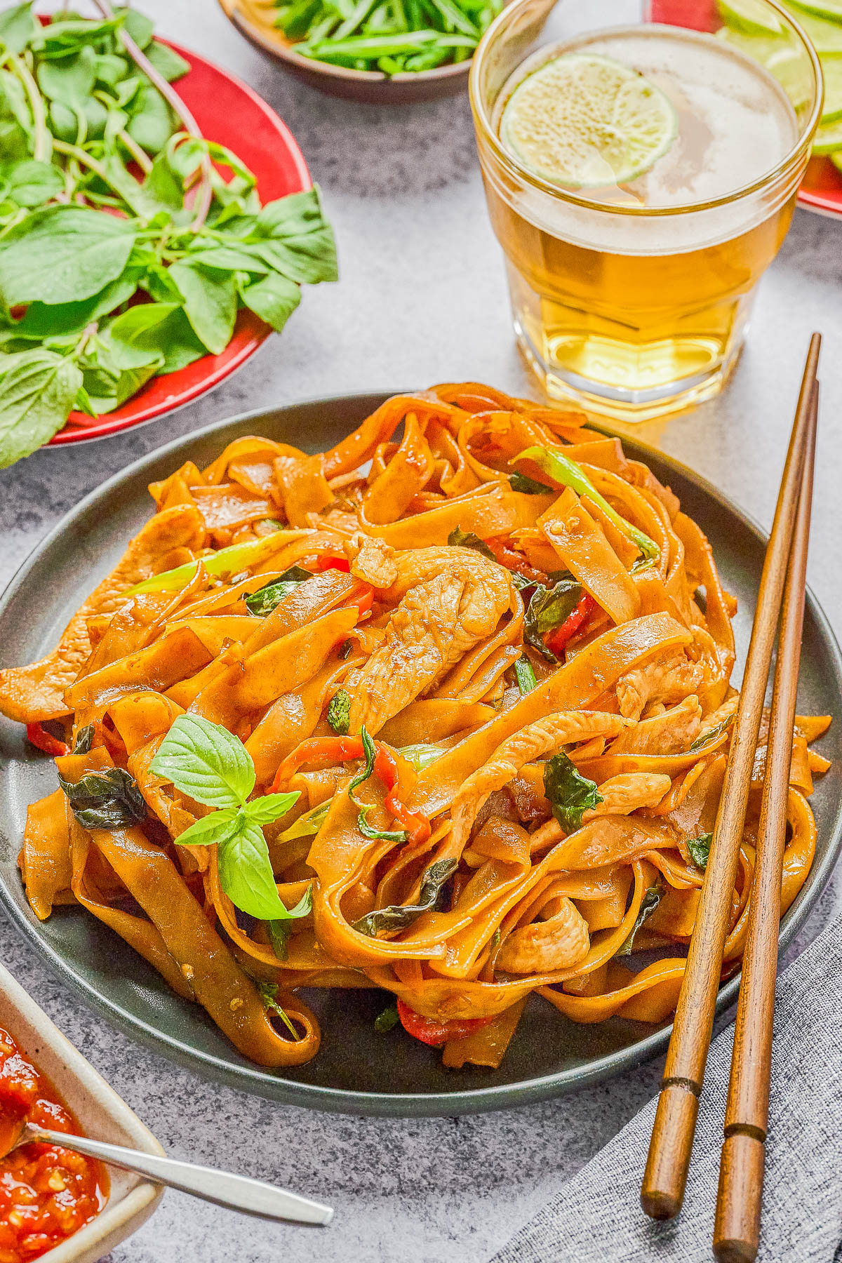 A plate of stir-fried noodles with vegetables and meat, garnished with basil, next to chopsticks, iced drink, and a side of fresh herbs.