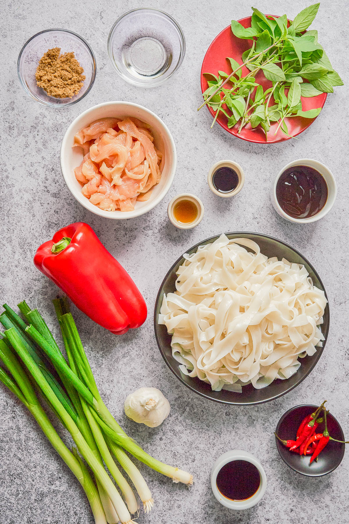 Various ingredients for a noodle dish are laid out, including rice noodles, raw chicken, red pepper, green onions, garlic, basil, chili, sauces, and seasonings on a gray surface.