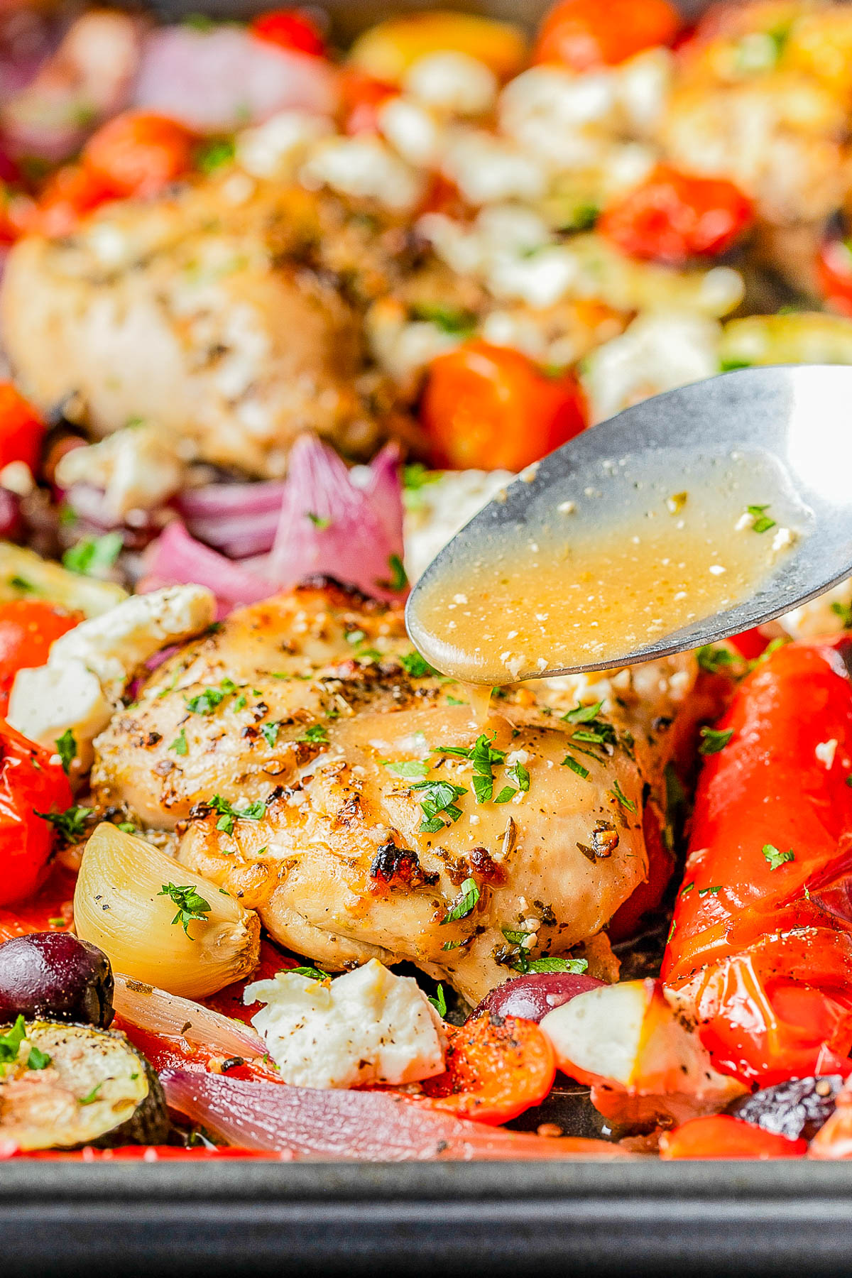 A spoon drizzles sauce over seasoned baked chicken surrounded by roasted vegetables, tomatoes, onions, and herbs on a sheet pan.