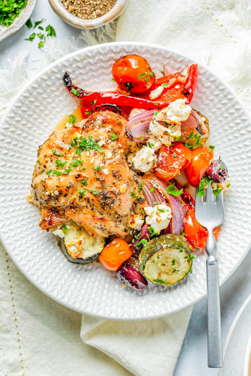 A plate of baked chicken with herbs, served with roasted zucchini, cherry tomatoes, red onion, red bell pepper, olives, and crumbled feta cheese. A fork rests on the plate.