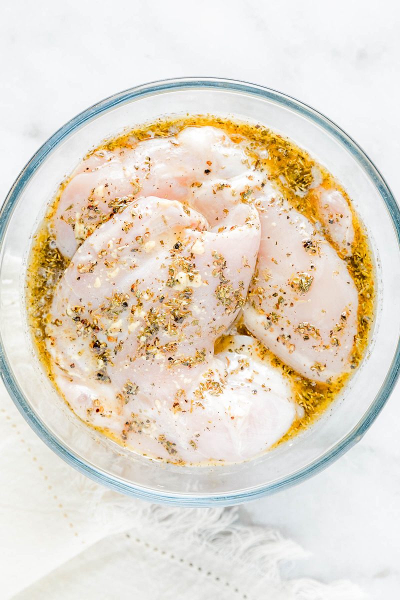 Raw chicken breasts marinating in a glass bowl with a mixture of oil, herbs, and spices on a white surface.