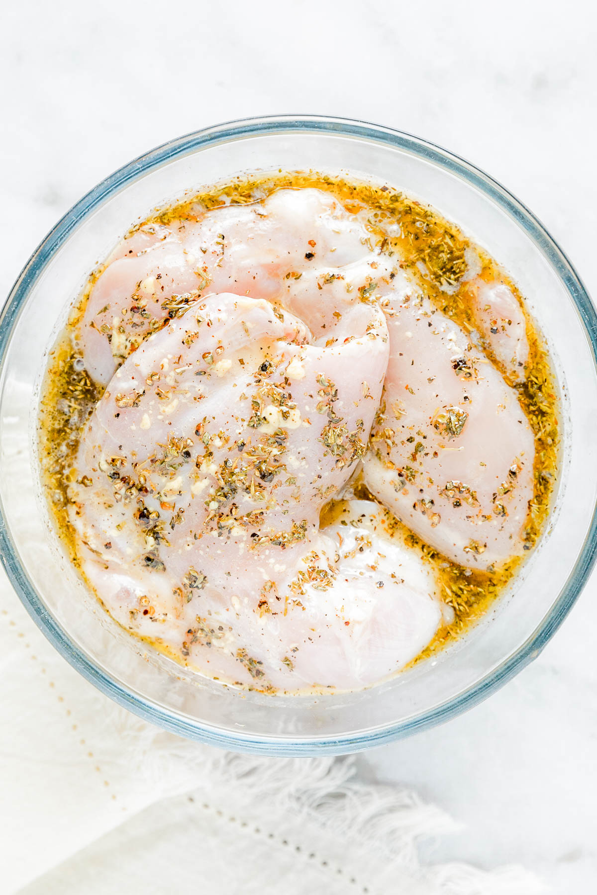 Raw chicken breasts marinating in a glass bowl with a mixture of oil, herbs, and spices on a white surface.