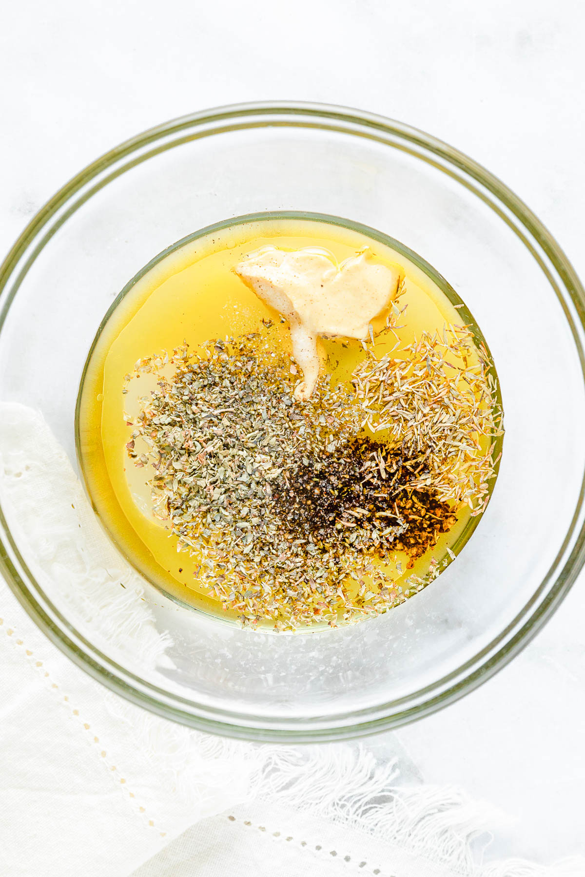 A glass bowl containing olive oil, Dijon mustard, dried herbs, and black pepper on a white surface.