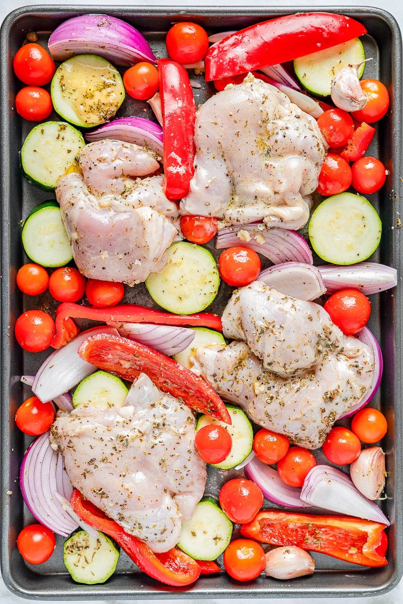 Raw chicken thighs, sliced zucchini, red onion, bell pepper, cherry tomatoes, and garlic cloves seasoned with herbs on a baking sheet.
