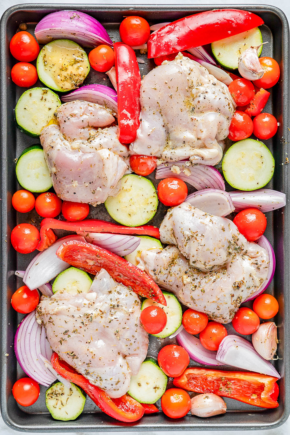 Raw chicken thighs, sliced zucchini, red onion, bell pepper, cherry tomatoes, and garlic cloves seasoned with herbs on a baking sheet.