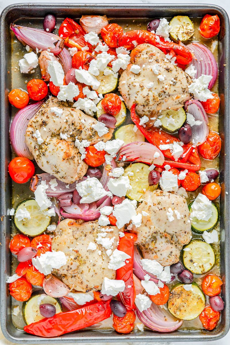 Sheet pan with baked chicken thighs, cherry tomatoes, zucchini, red onion, red bell pepper, Kalamata olives, and crumbled feta cheese.