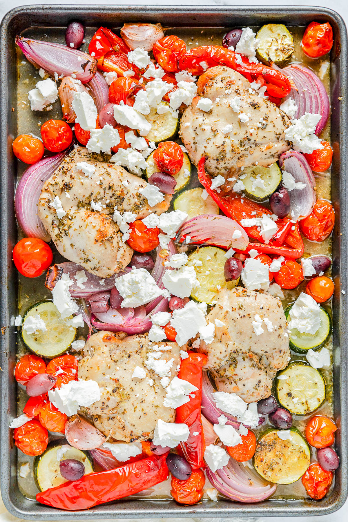 Sheet pan with baked chicken thighs, cherry tomatoes, zucchini, red onion, red bell pepper, Kalamata olives, and crumbled feta cheese.