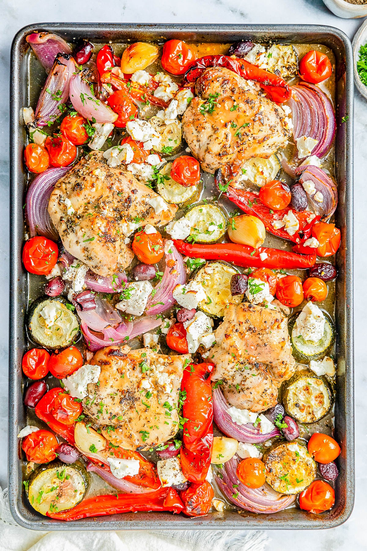 Sheet pan with baked chicken thighs, red onions, zucchini, cherry tomatoes, red bell peppers, Kalamata olives, and crumbled feta cheese, garnished with herbs.
