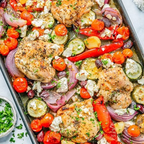 Baked chicken thighs with cherry tomatoes, zucchini, red onions, red bell peppers, olives, and crumbled feta on a sheet pan, garnished with chopped parsley.