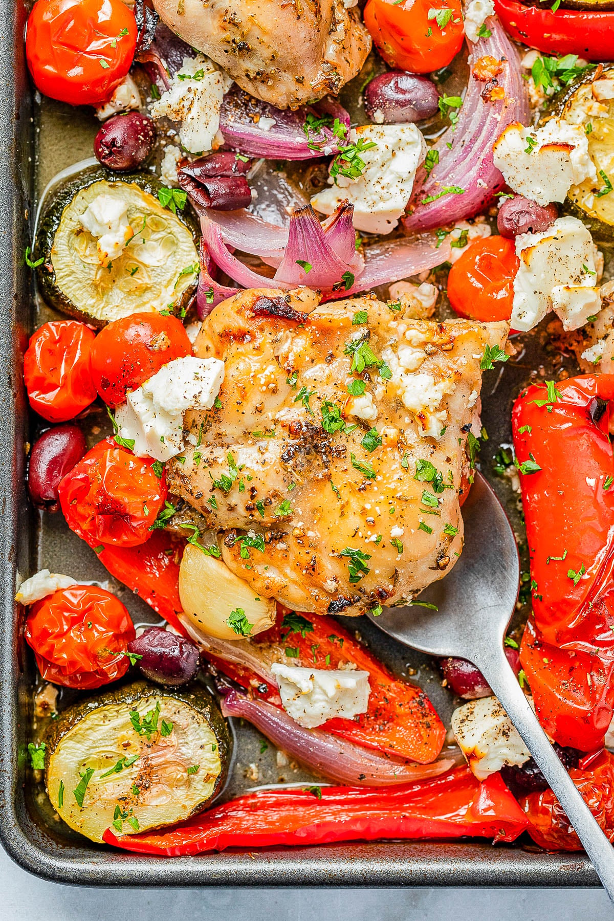 A sheet pan with baked chicken, cherry tomatoes, red peppers, zucchini, red onions, olives, and crumbled cheese, garnished with chopped herbs.