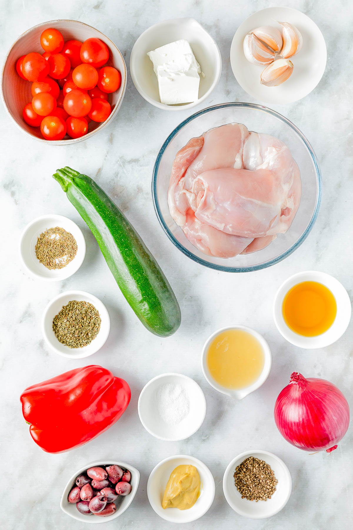 Ingredients arranged on a white surface, including raw chicken, cherry tomatoes, zucchini, red pepper, onion, garlic, feta, olives, and various spices and condiments in small bowls.