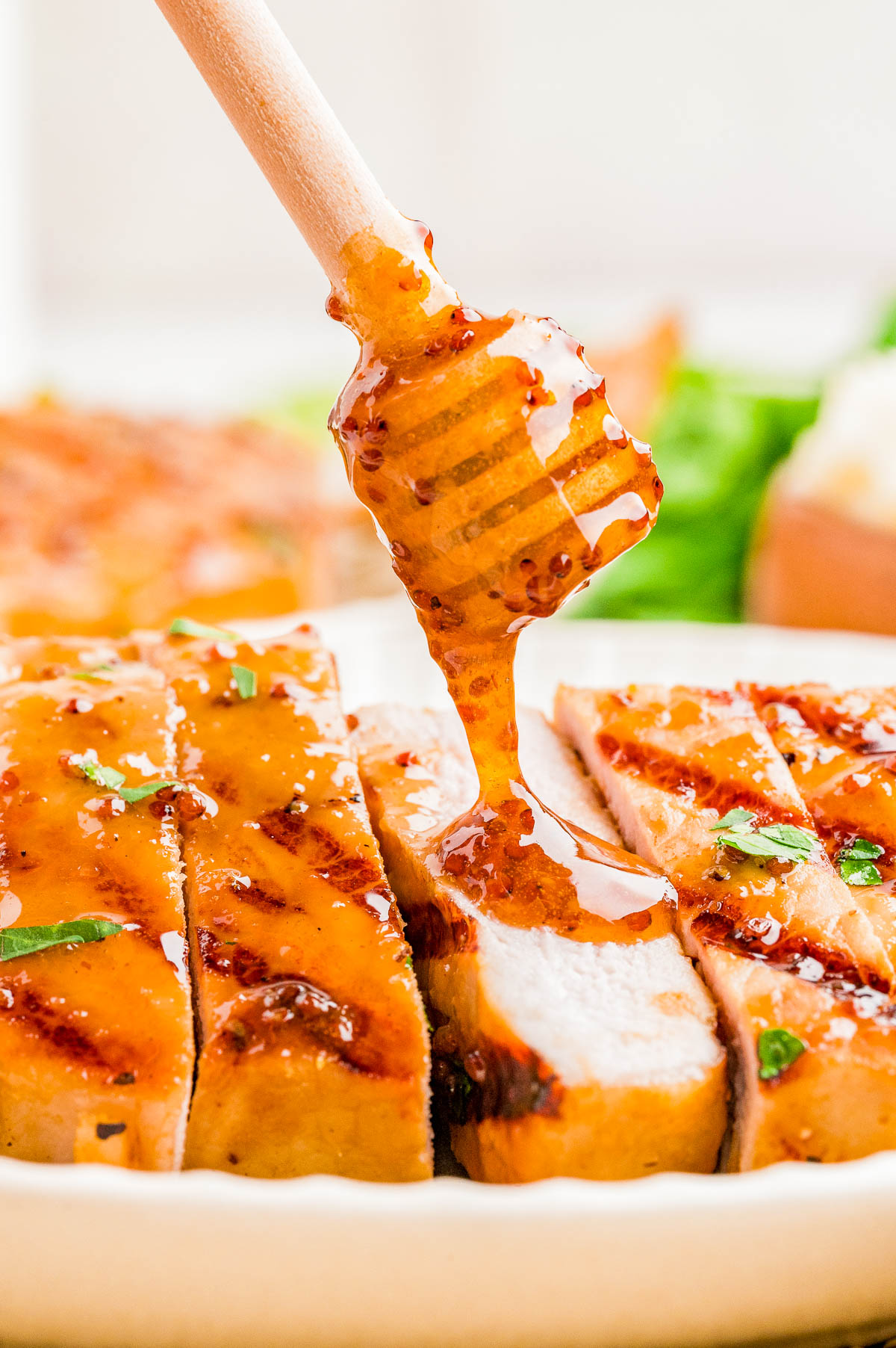 Close-up of sliced grilled chicken breast being drizzled with honey mustard sauce using a wooden honey dipper.