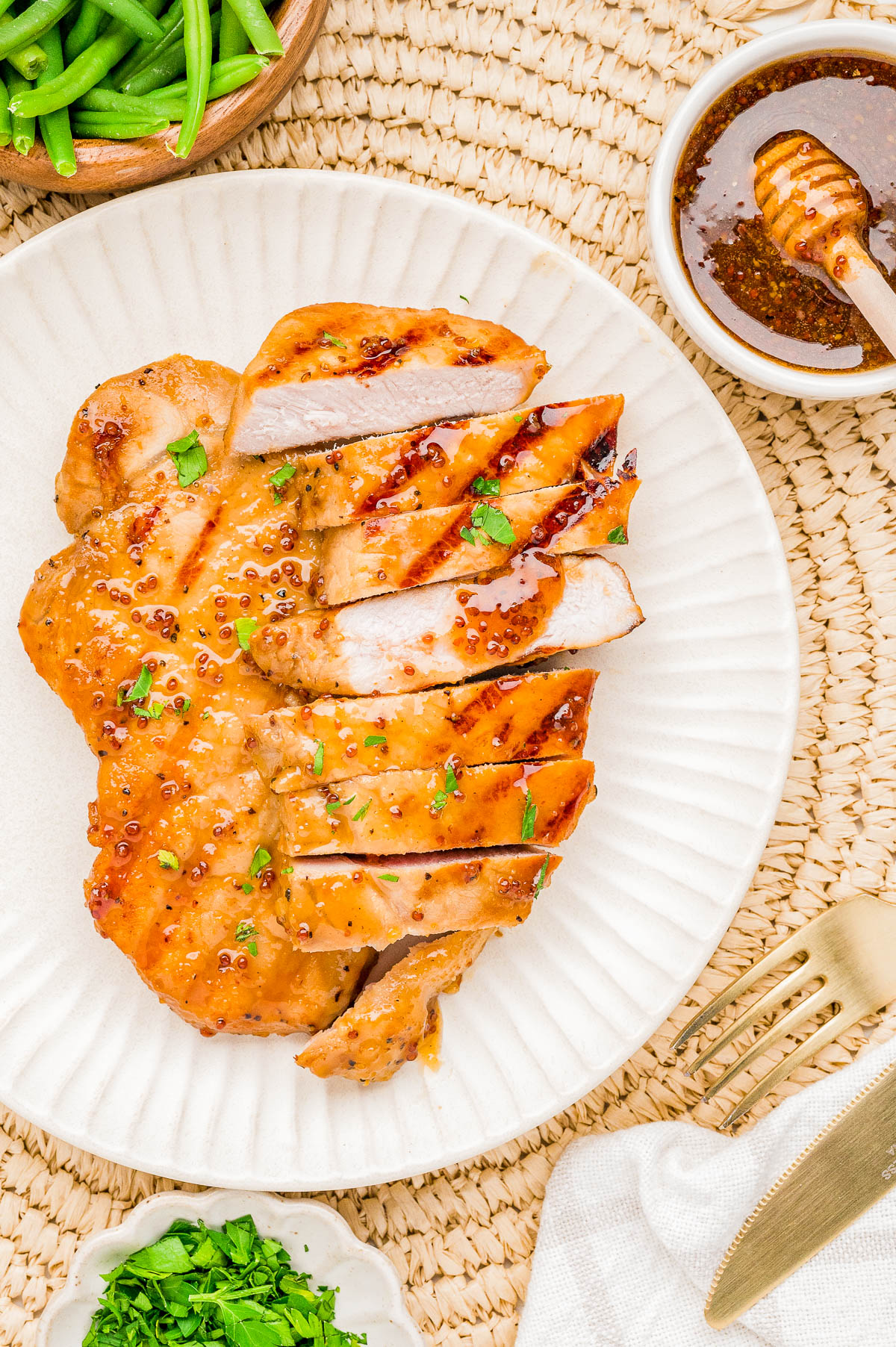 Sliced grilled chicken breast with a honey mustard glaze, garnished with herbs, served on a white plate with sides of green beans and chopped herbs.