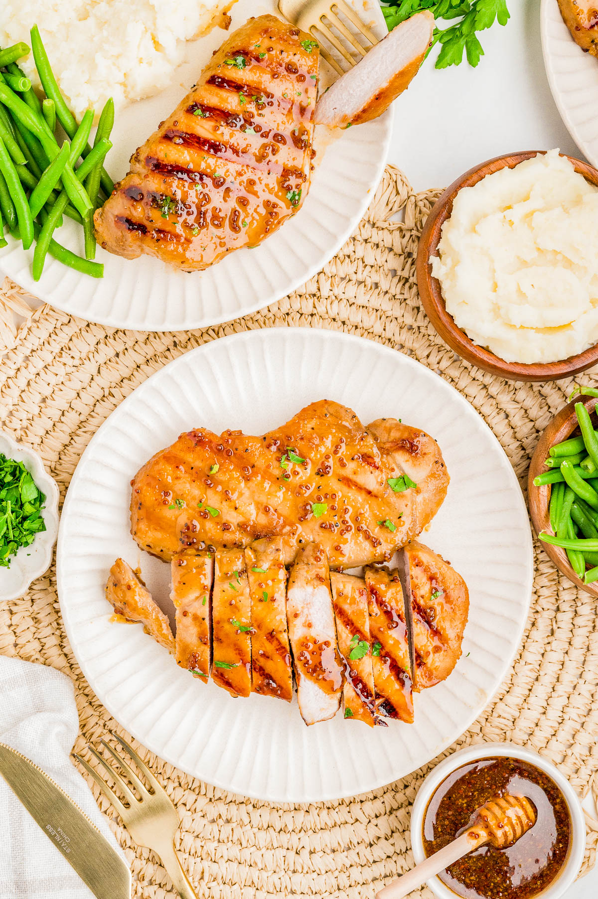 Grilled pork chops served with green beans and mashed potatoes on white plates, accompanied by a small bowl of sauce and fresh herbs on a woven placemat.