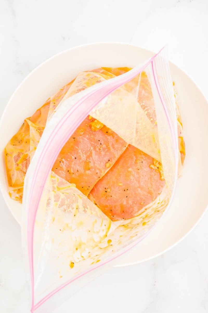 Raw chicken breasts marinating in a sealed plastic zip-top bag placed inside a white bowl on a white surface.