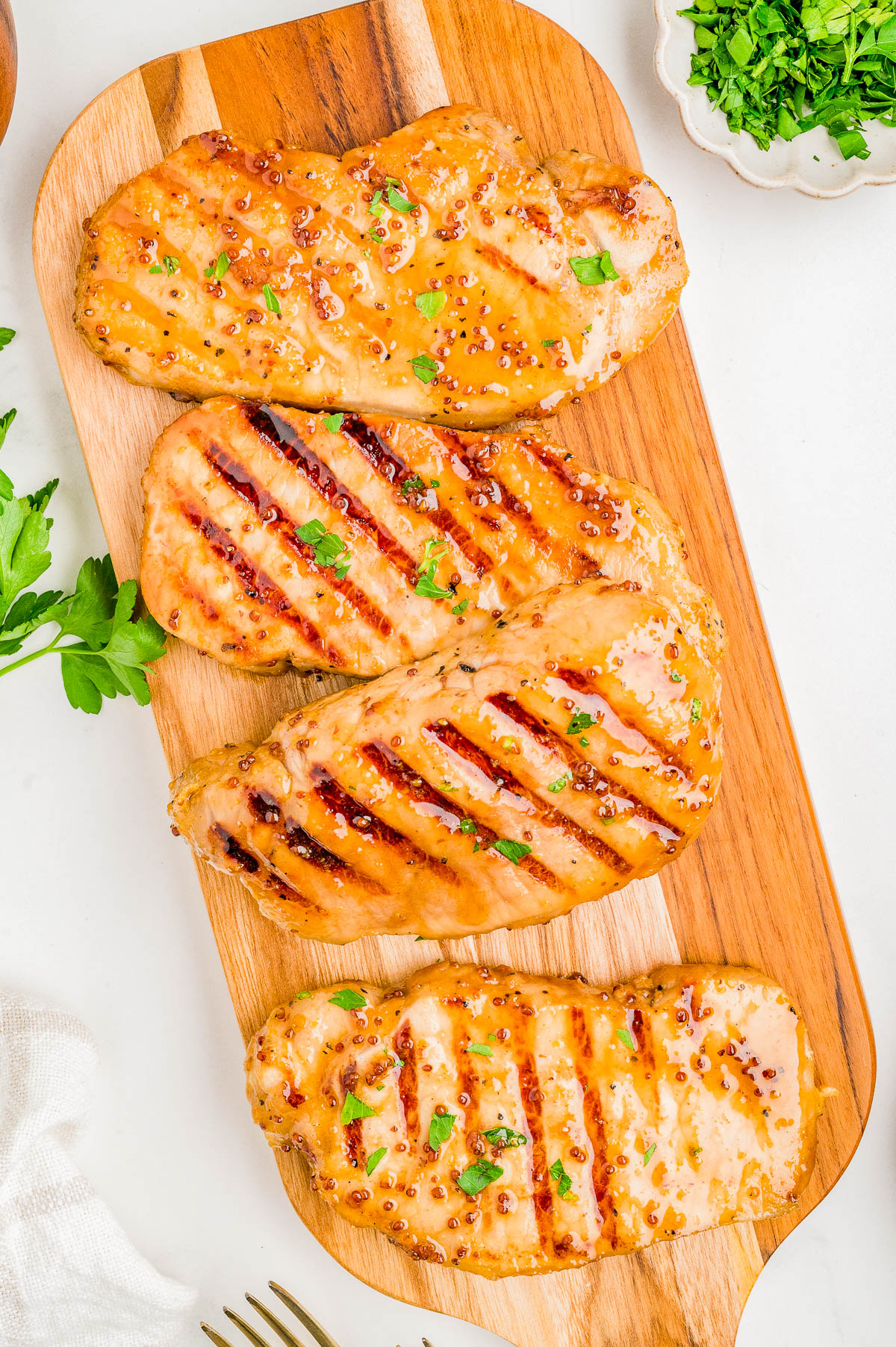 Four grilled chicken breasts with visible grill marks and a glaze, garnished with parsley, are arranged on a wooden cutting board.