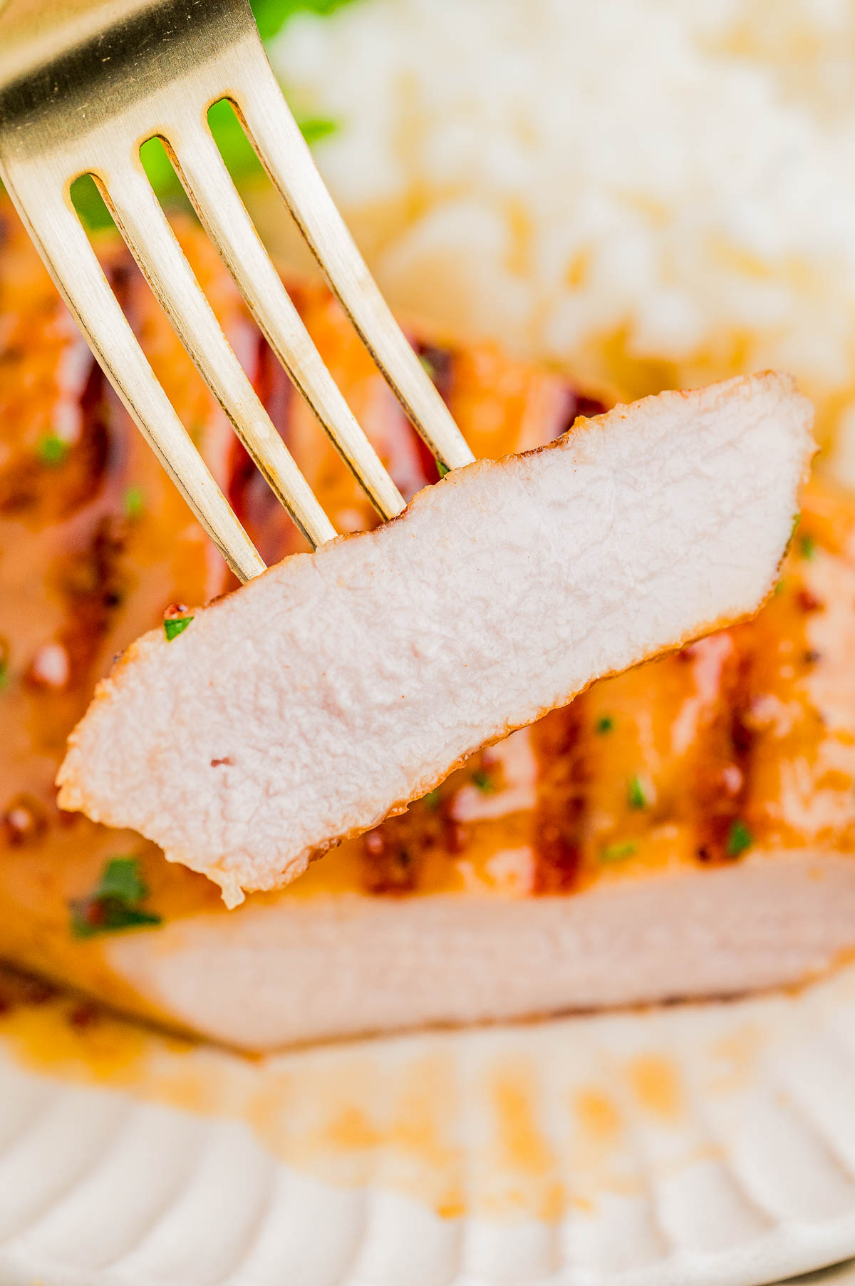A close-up of a fork holding a slice of grilled pork chop, with grill marks visible and rice in the background.
