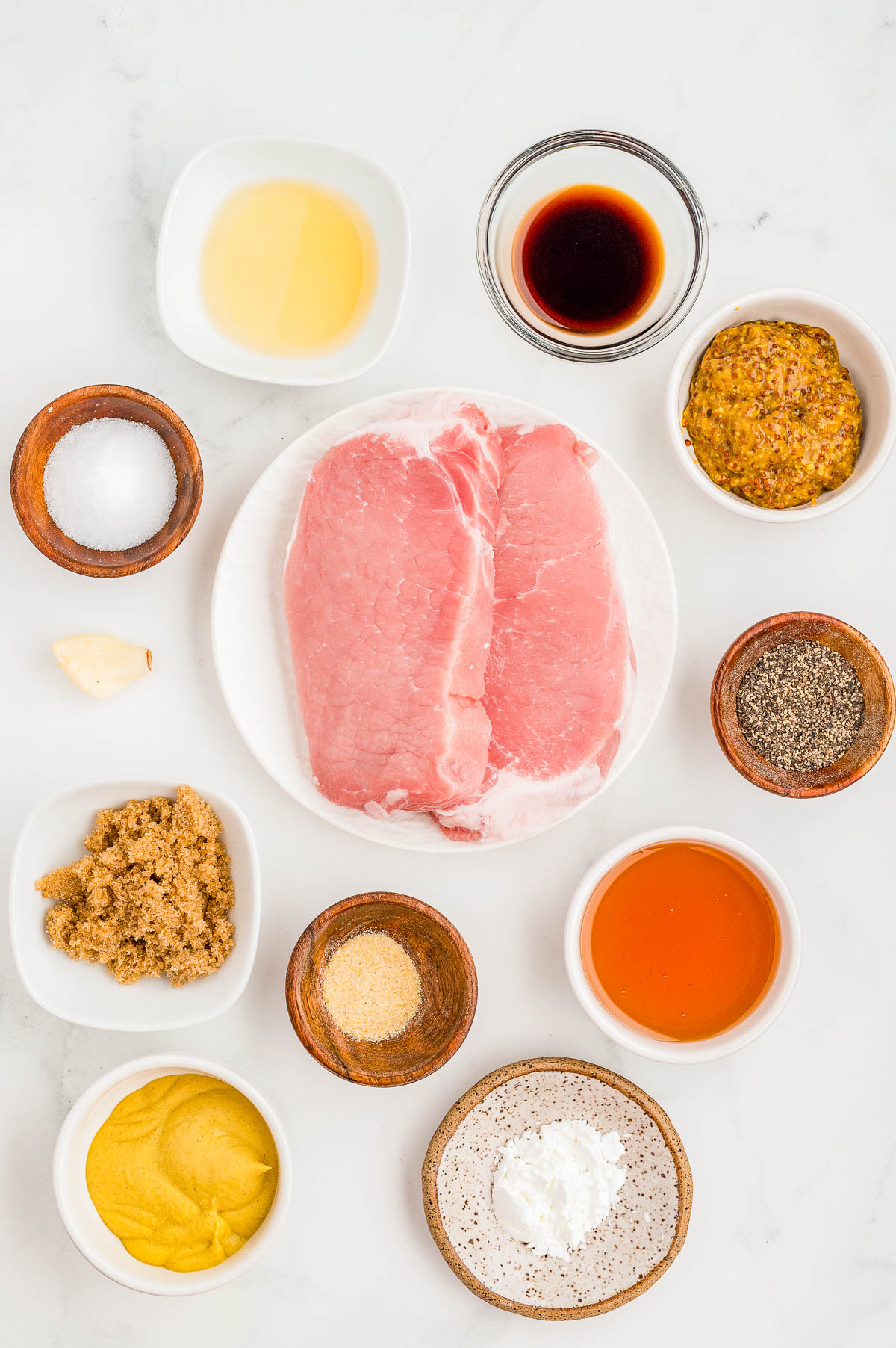 Two raw pork chops on a plate, surrounded by small bowls of mustard, spices, brown sugar, vinegar, soy sauce, honey, cornstarch, and a garlic clove on a white surface.