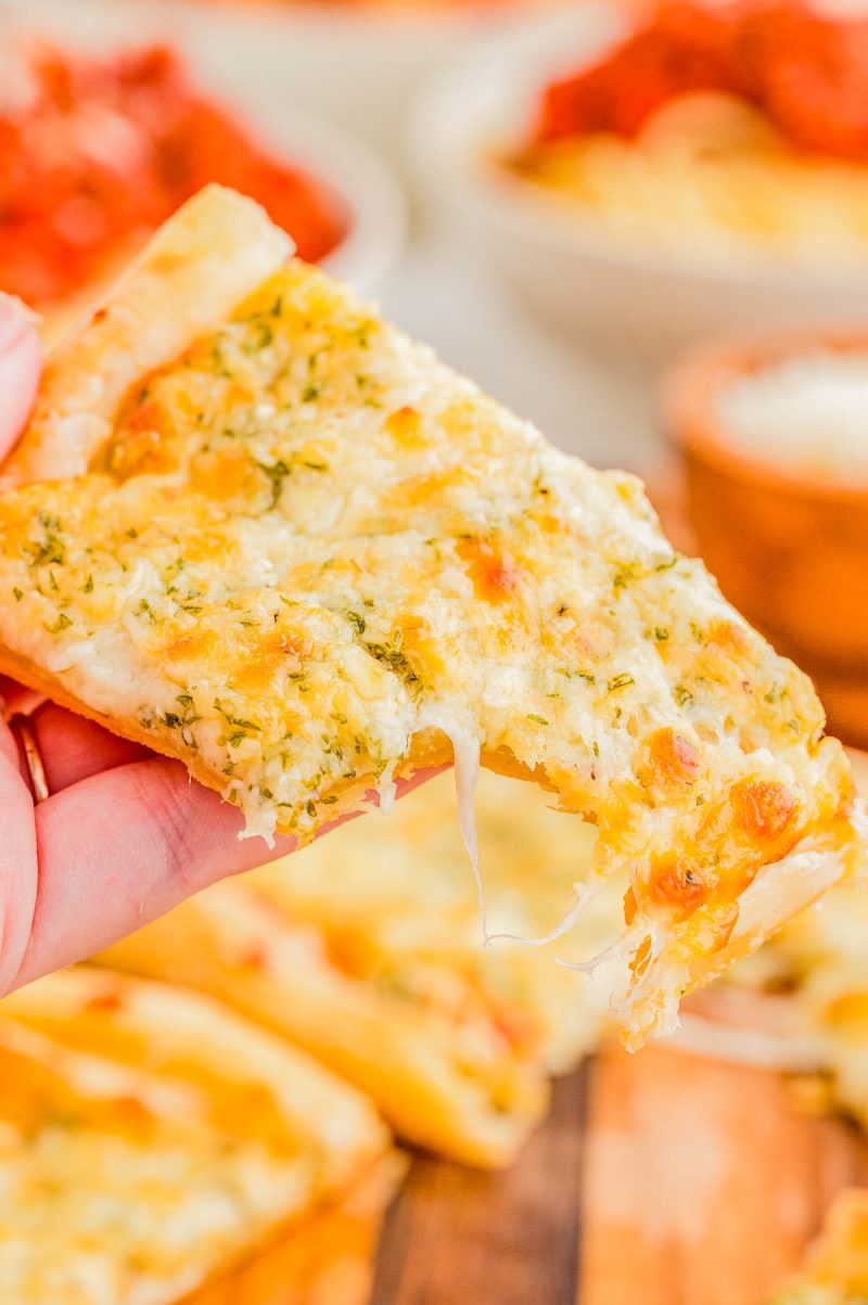 A hand holds a slice of cheesy garlic bread with herbs, showing melted cheese stretching from the crust.