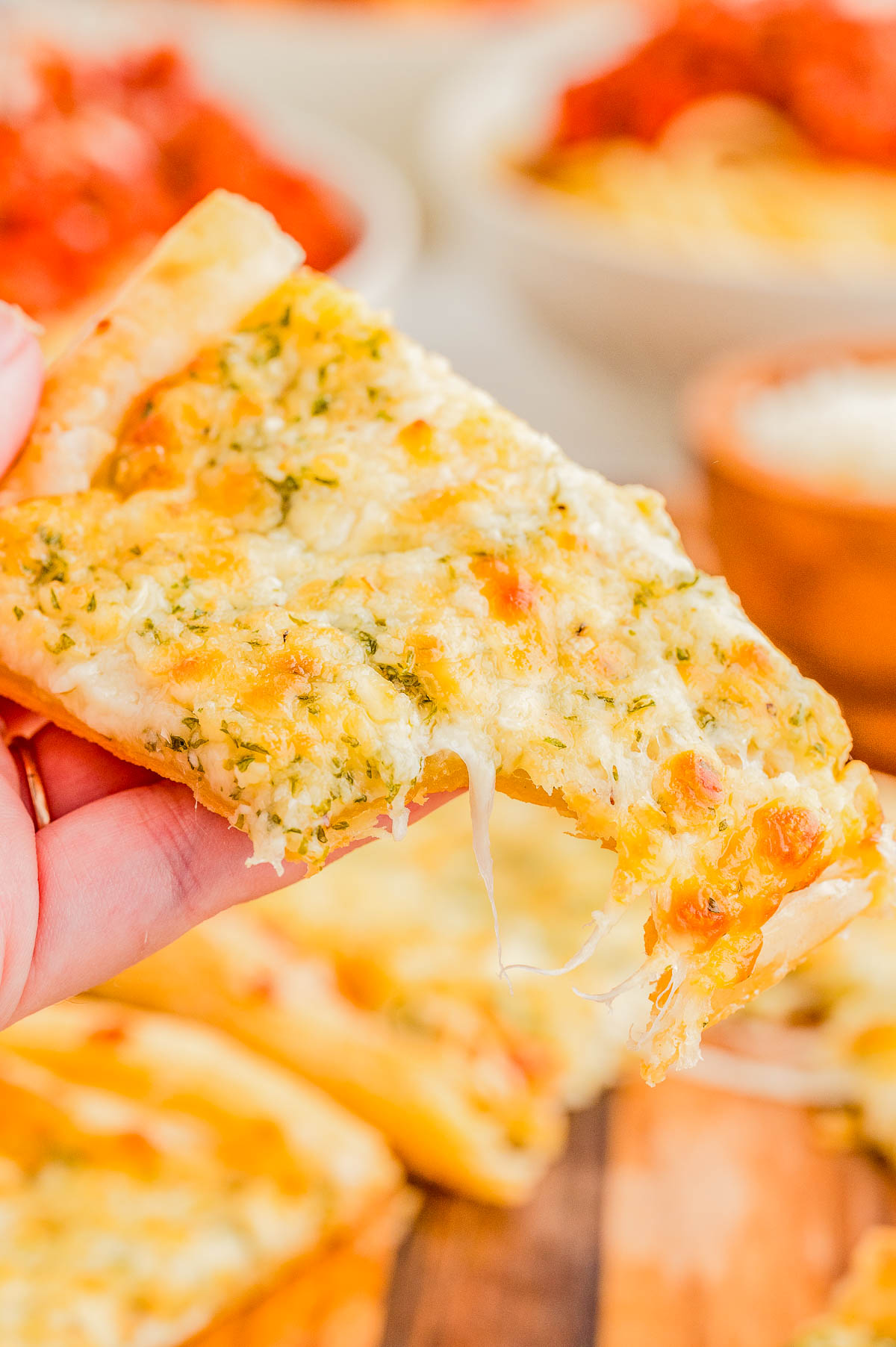 A hand holds a slice of cheesy garlic bread with herbs, showing melted cheese stretching from the crust.