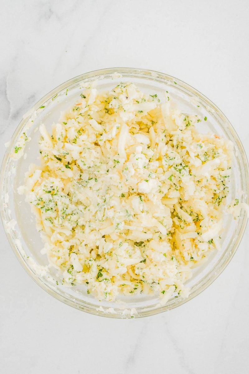 A glass bowl filled with a mixture of shredded potatoes, cheese, herbs, and a creamy dressing on a white surface.