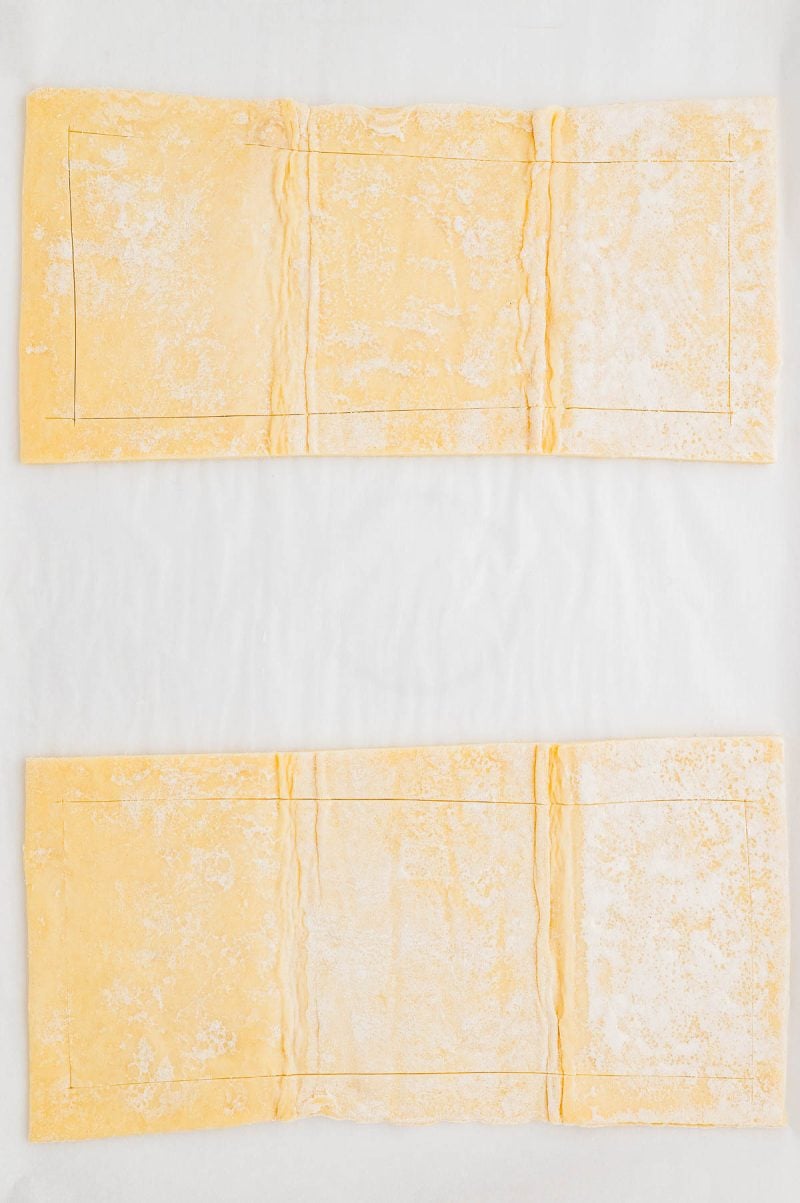 Two rectangular sheets of uncooked puff pastry are laid out flat on a white surface, with faint lines marking smaller rectangles on each sheet.