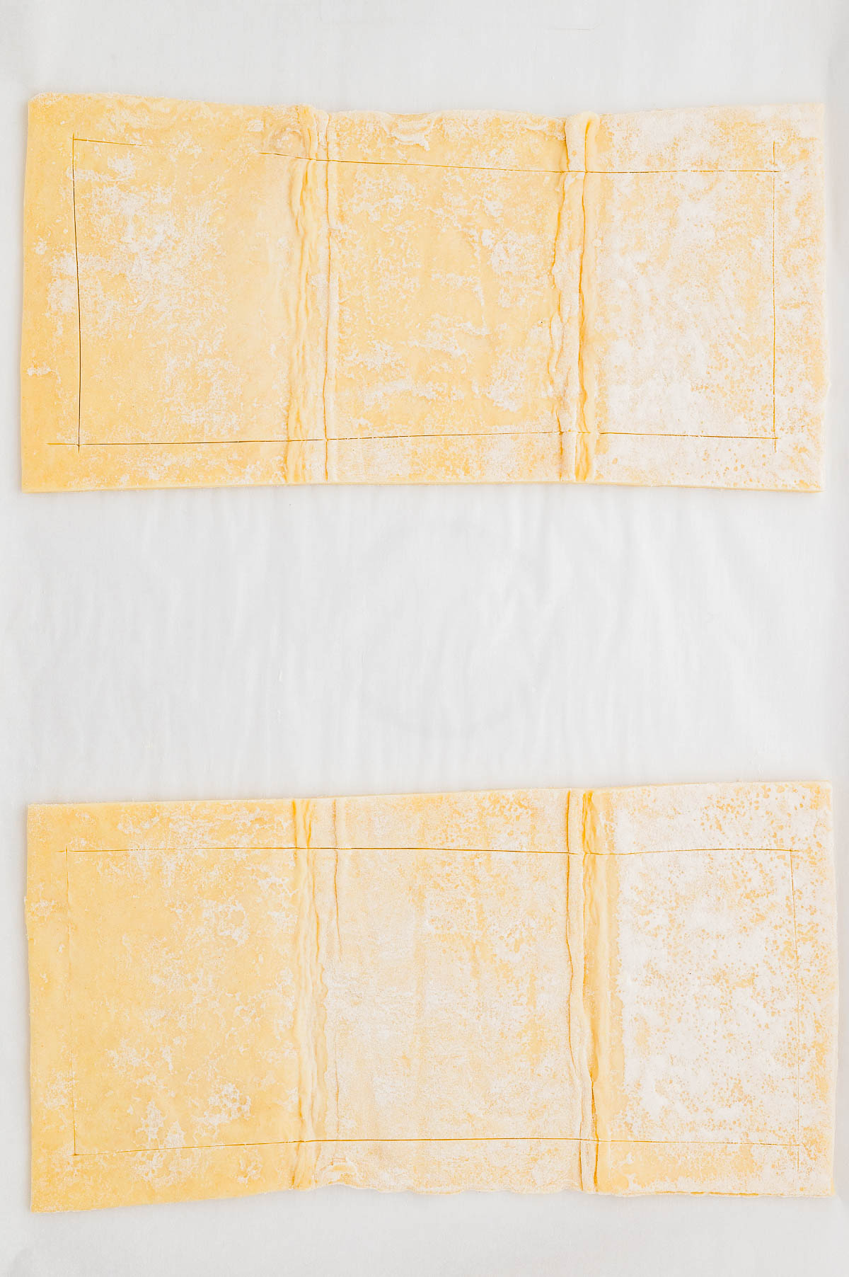 Two rectangular sheets of uncooked puff pastry are laid out flat on a white surface, with faint lines marking smaller rectangles on each sheet.