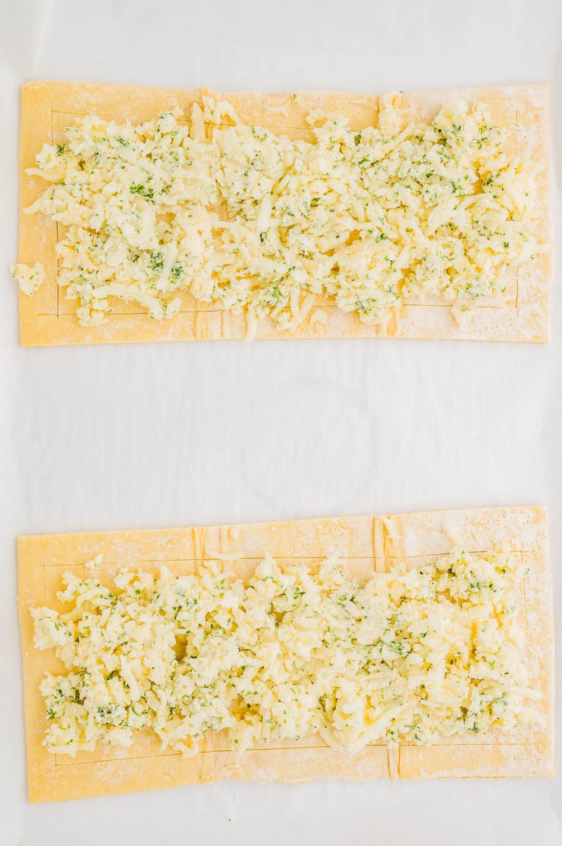 Two rectangular sheets of dough topped with a mixture of shredded cheese and herbs, laid out on a white surface.