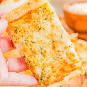 A hand holds a rectangular slice of cheesy garlic bread over a wooden surface, with more slices and bowls of food visible in the background.