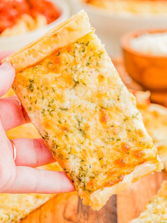 A hand holds a rectangular slice of cheesy garlic bread over a wooden surface, with more slices and bowls of food visible in the background.
