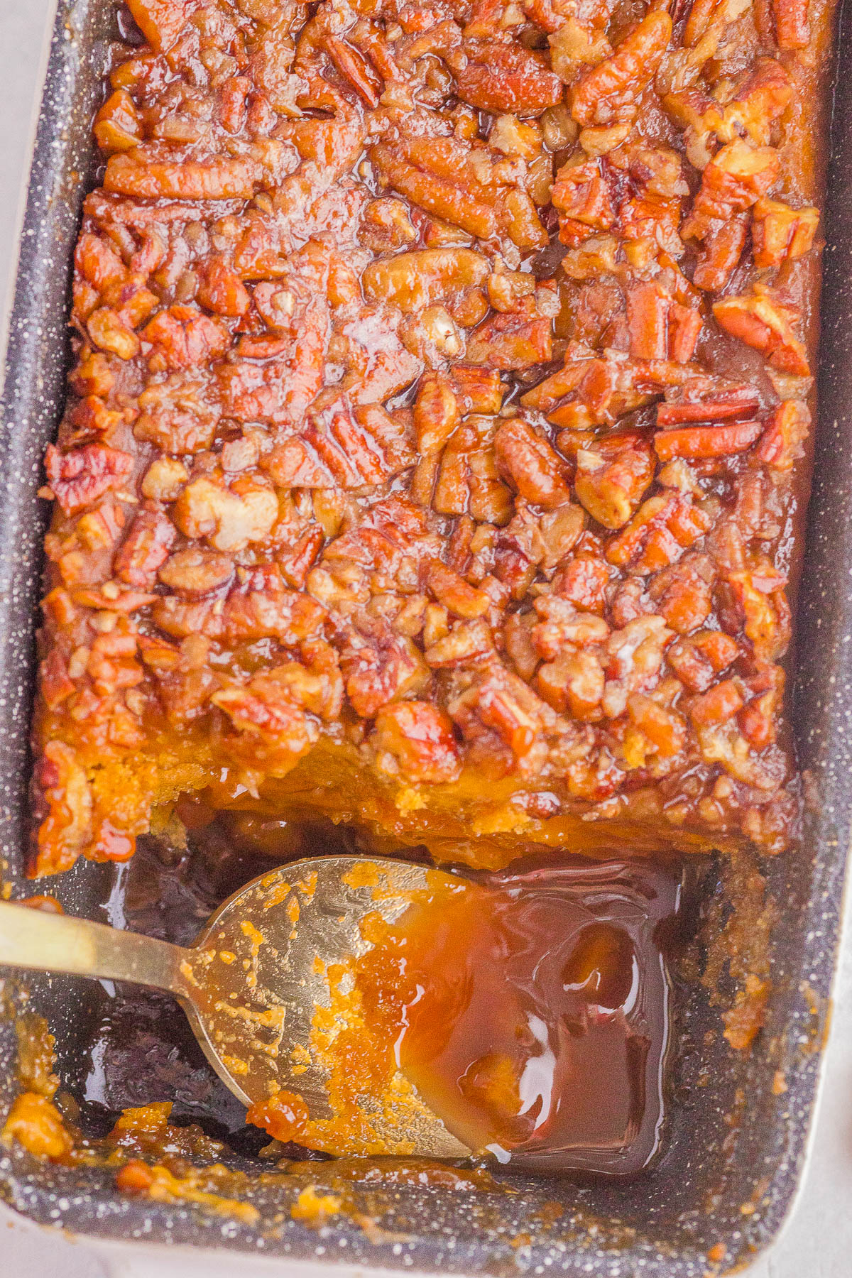 A close-up of a baked pecan-topped dessert in a pan with a portion removed, revealing a gooey caramel sauce and a spoon resting inside.