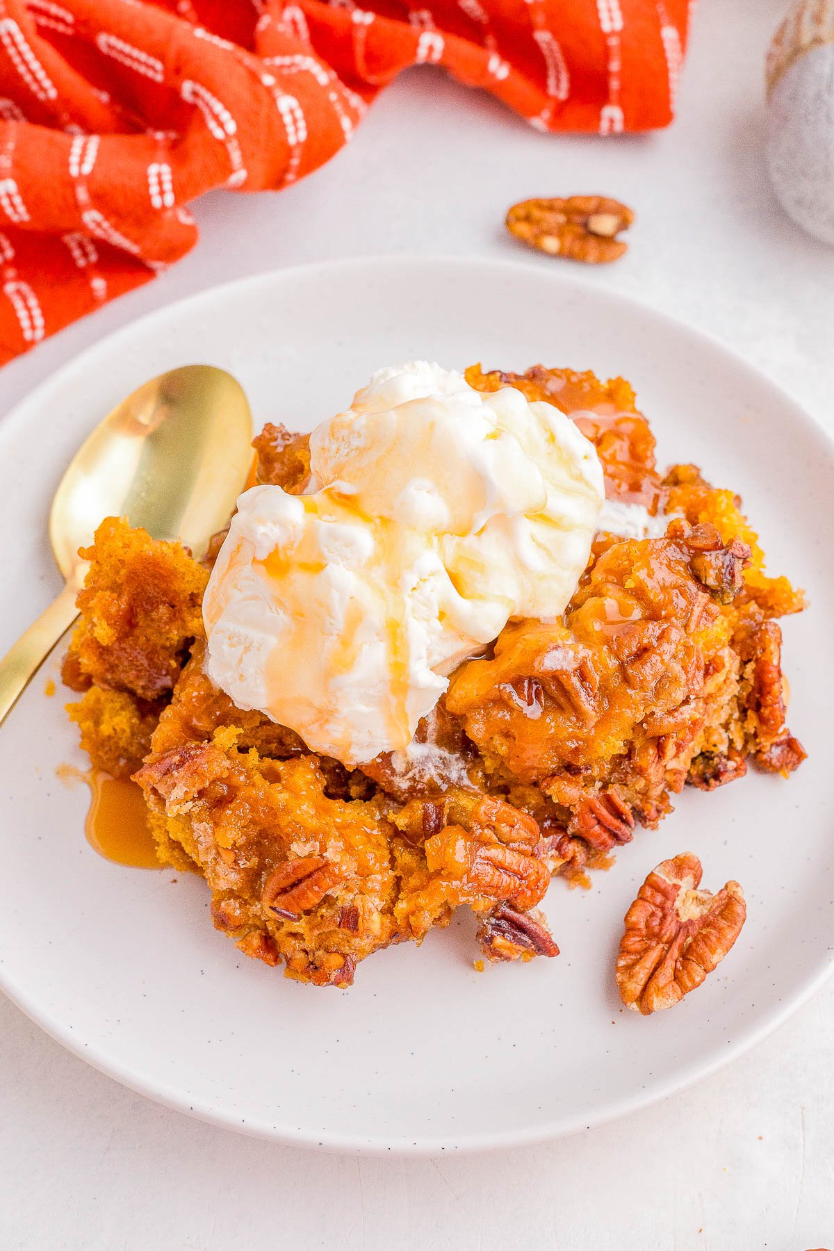 A serving of pecan pumpkin dessert topped with a scoop of vanilla ice cream and caramel sauce on a white plate with a gold spoon and pecans.